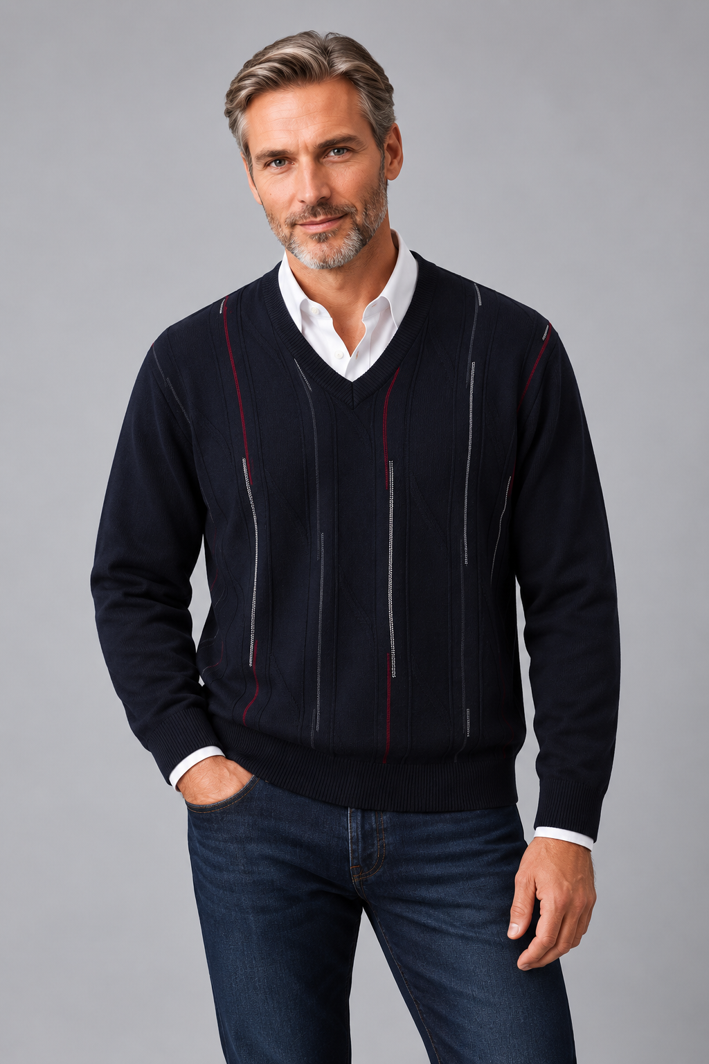 A man wearing a dark navy blue V-neck sweater with vertical knit stripe details in red, white, and gray, layered over a white collared shirt and paired with dark blue jeans, against a solid gray studio background.