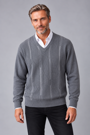 A man wearing a gray v-neck sweater with vertical knit stripe details over a white collared shirt and black pants, photographed against a solid gray studio background.