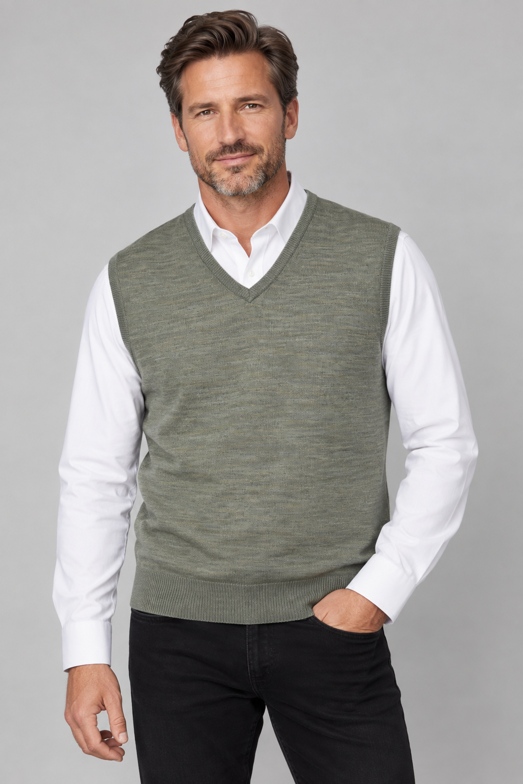 A man wearing a sage green V-neck knit sweater vest over a white collared shirt and black pants, standing against a light gray studio background.