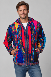A man wearing a vibrant, multicolored hooded jacket with intricate geometric and tribal patterns in red, blue, yellow, and black, paired with blue jeans against a solid gray studio background.