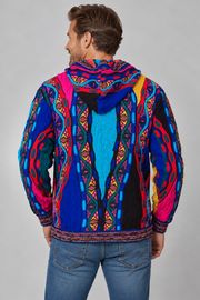 A man viewed from the back wearing a vibrant multicolor hooded jacket with intricate geometric and tribal patterns in blue, pink, red, yellow, and black, paired with blue jeans against a solid gray studio background.
