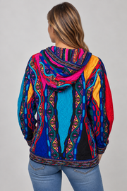 A woman viewed from the back wearing a vibrant, multicolored hooded sweatshirt with intricate geometric and tribal-inspired patterns in blue, pink, red, yellow, and black, paired with blue jeans against a neutral gray background.