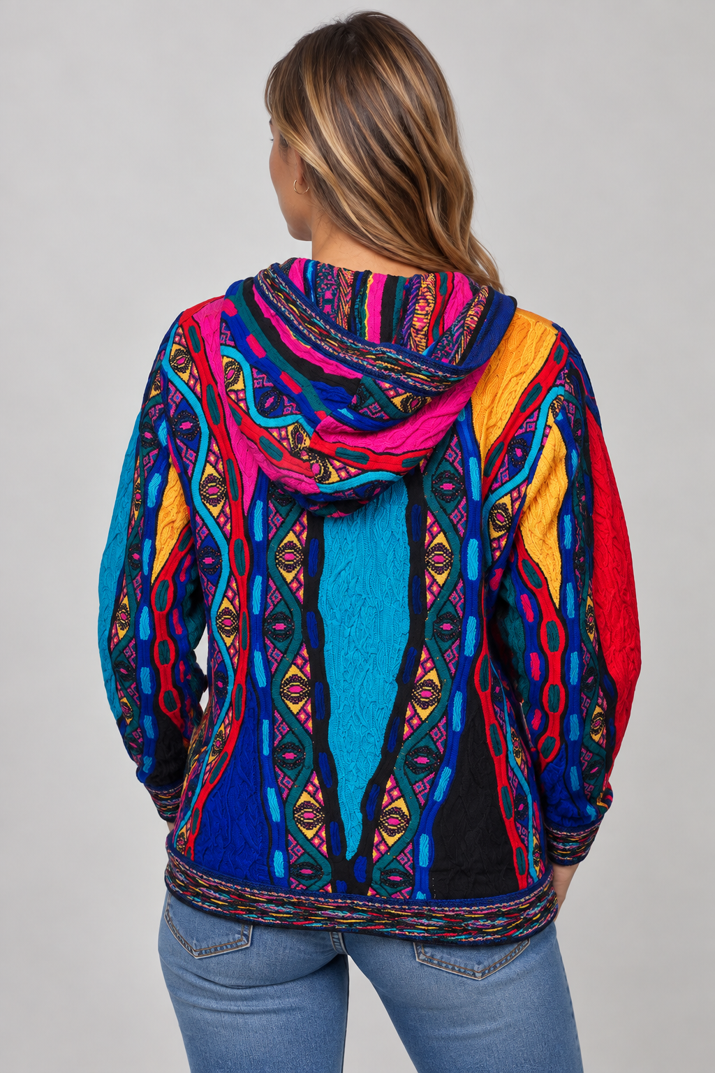A woman viewed from the back wearing a vibrant, multicolored hooded sweatshirt with intricate geometric and tribal-inspired patterns in blue, pink, red, yellow, and black, paired with blue jeans against a neutral gray background.