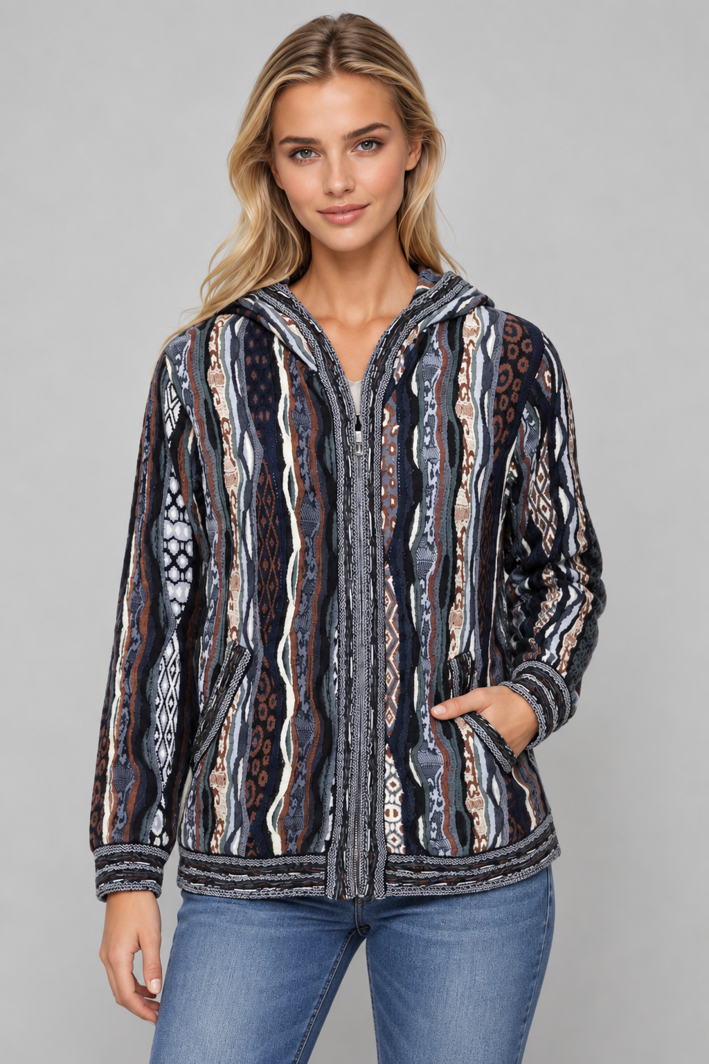 A woman modeling a zip-up hooded jacket with a vertical tribal pattern in navy, beige, brown, and white, paired with blue jeans against a solid gray studio background.