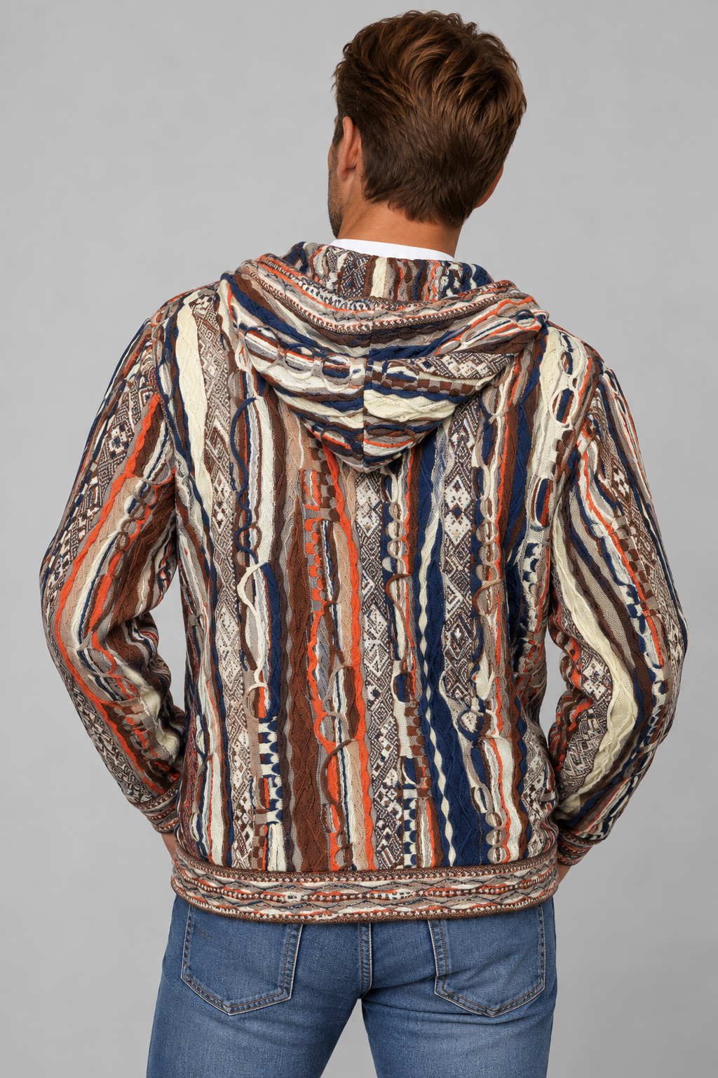 A man viewed from behind wearing a hooded jacket with a complex vertical tribal-style pattern in beige, brown, orange, and navy blue, paired with blue jeans against a solid gray background.