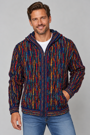 A man wearing a colorful zip-up hooded jacket with a vertical multicolor pattern in red, blue, yellow, and orange over a white t-shirt and blue jeans, photographed against a solid gray studio background.