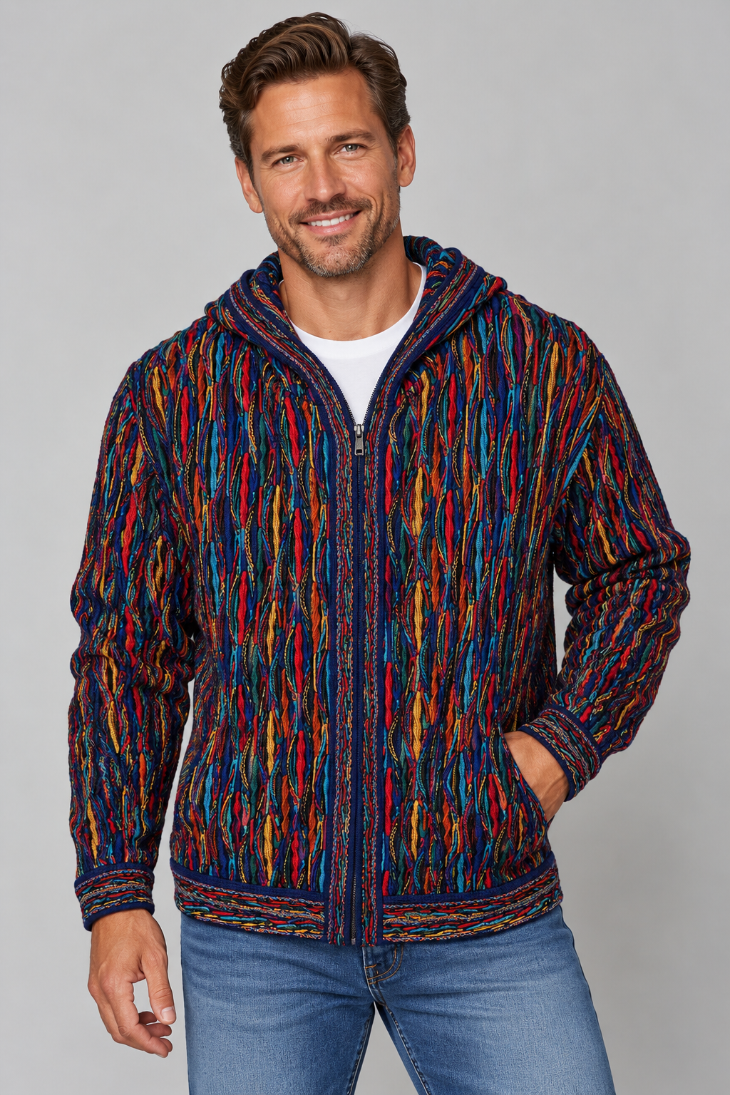A man wearing a colorful zip-up hooded jacket with a vertical multicolor pattern in red, blue, yellow, and orange over a white t-shirt and blue jeans, photographed against a solid gray studio background.