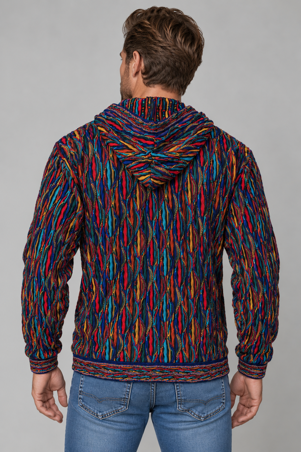 A man viewed from behind wearing a hooded sweatshirt with a vibrant multicolor abstract pattern in red, blue, yellow, and orange, paired with blue jeans against a solid gray studio background.