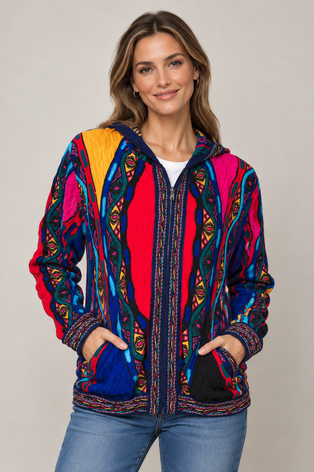 A woman wearing a vibrant, multicolored hooded jacket with intricate geometric and tribal patterns in red, blue, pink, yellow, and black, paired with blue jeans, against a neutral gray studio background.