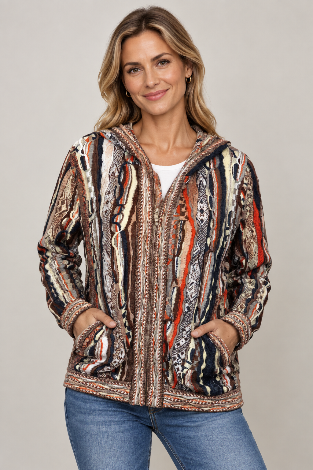 A woman modeling a hooded zip-up jacket with a complex vertical tribal pattern in brown, beige, black, white, and red, paired with blue jeans against a neutral gray background.