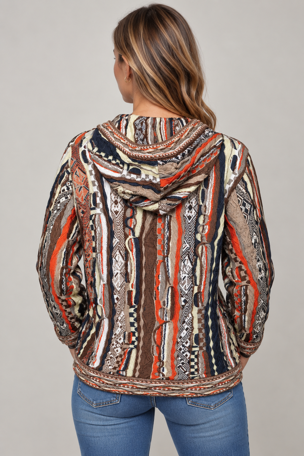 A woman viewed from the back wearing a hooded sweatshirt with a complex vertical tribal-style pattern in orange, brown, black, beige, and white, paired with blue jeans against a solid gray background.