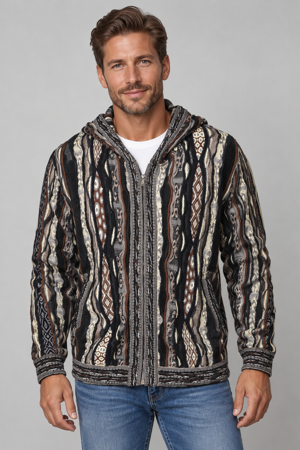 A man wearing a black, brown, and beige patterned zip-up hooded jacket over a white t-shirt and blue jeans, standing against a solid gray studio background.
