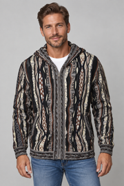 A man wearing a black, brown, and beige patterned zip-up hooded jacket over a white t-shirt and blue jeans, standing against a solid gray studio background.