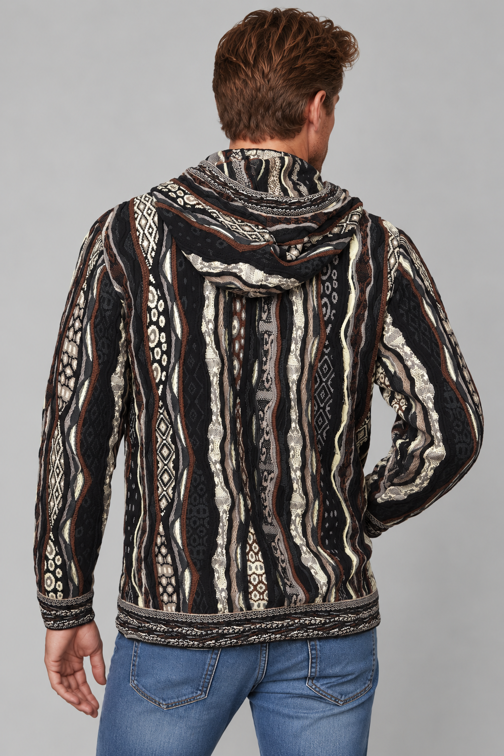 A man viewed from the back wearing a hooded long-sleeve shirt with a black, beige, and brown tribal-style geometric pattern, paired with blue jeans, against a solid gray background.