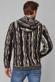A man viewed from the back wearing a hooded long-sleeve shirt with a black, beige, and brown tribal-style geometric pattern, paired with blue jeans, against a solid gray background.
