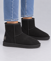 A pair of black suede ankle Ugg boots with black soles, shown on a model wearing blue jeans against a light grey background.