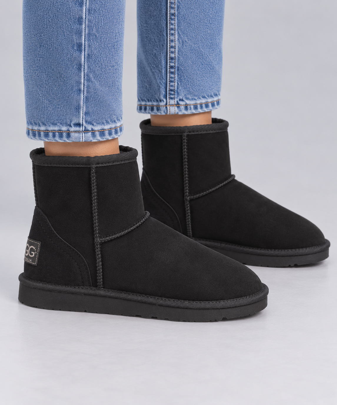 A pair of black suede ankle Ugg boots with black soles, shown on a model wearing blue jeans against a light grey background.