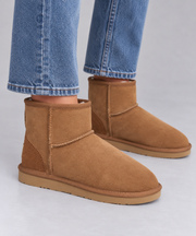 A pair of tan suede ankle ugg boots worn with blue jeans, shown from the side against a light grey studio background.