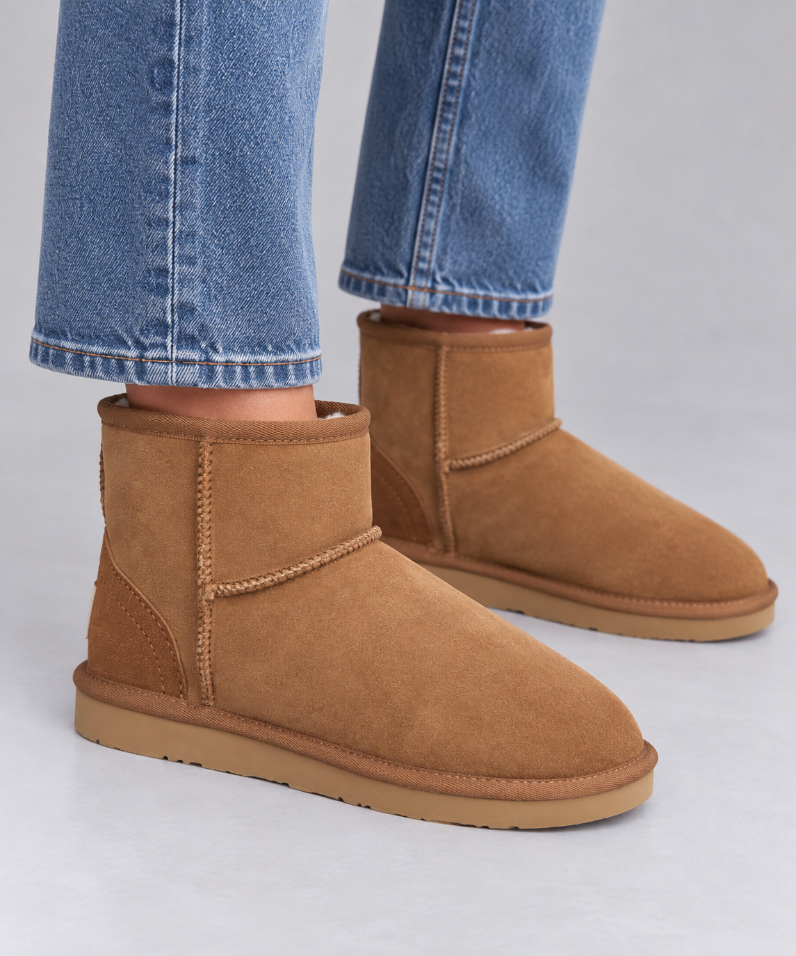 A pair of tan suede ankle ugg boots worn with blue jeans, shown from the side against a light grey studio background.