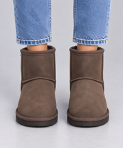 A pair of brown suede ankle Ugg boots with black soles, shown on a model wearing blue jeans against a light grey background.