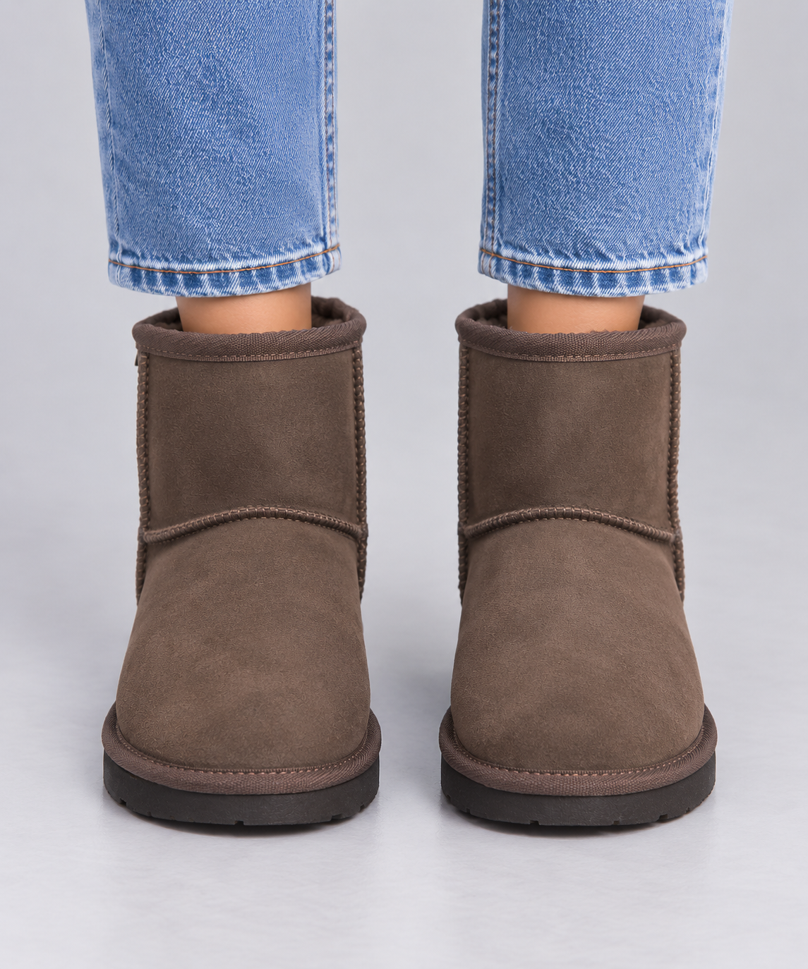 A pair of brown suede ankle Ugg boots with black soles, shown on a model wearing blue jeans against a light grey background.