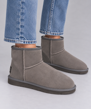 A pair of grey suede ankle ugg boots with grey trim and soles, shown on a model wearing blue jeans against a light grey studio background.