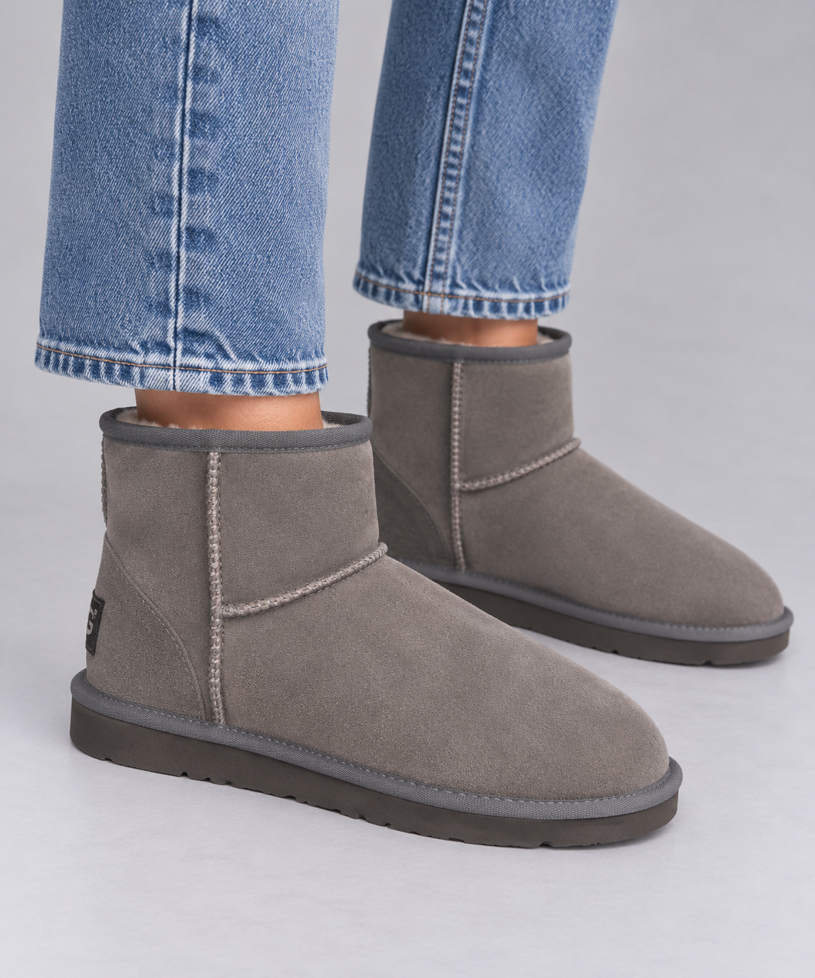 A pair of grey suede ankle ugg boots with grey trim and soles, shown on a model wearing blue jeans against a light grey studio background.