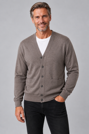 A smiling man wearing a taupe brown button-up cardigan over a white t-shirt and black jeans, standing against a solid gray studio background.