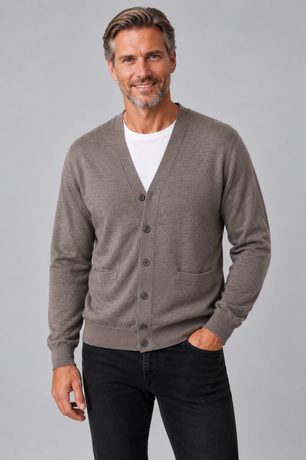 A smiling man wearing a taupe brown button-up cardigan over a white t-shirt and black jeans, standing against a solid gray studio background.