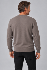A man viewed from the back wearing a taupe-gray crewneck sweater and black jeans, standing against a solid light gray studio background.