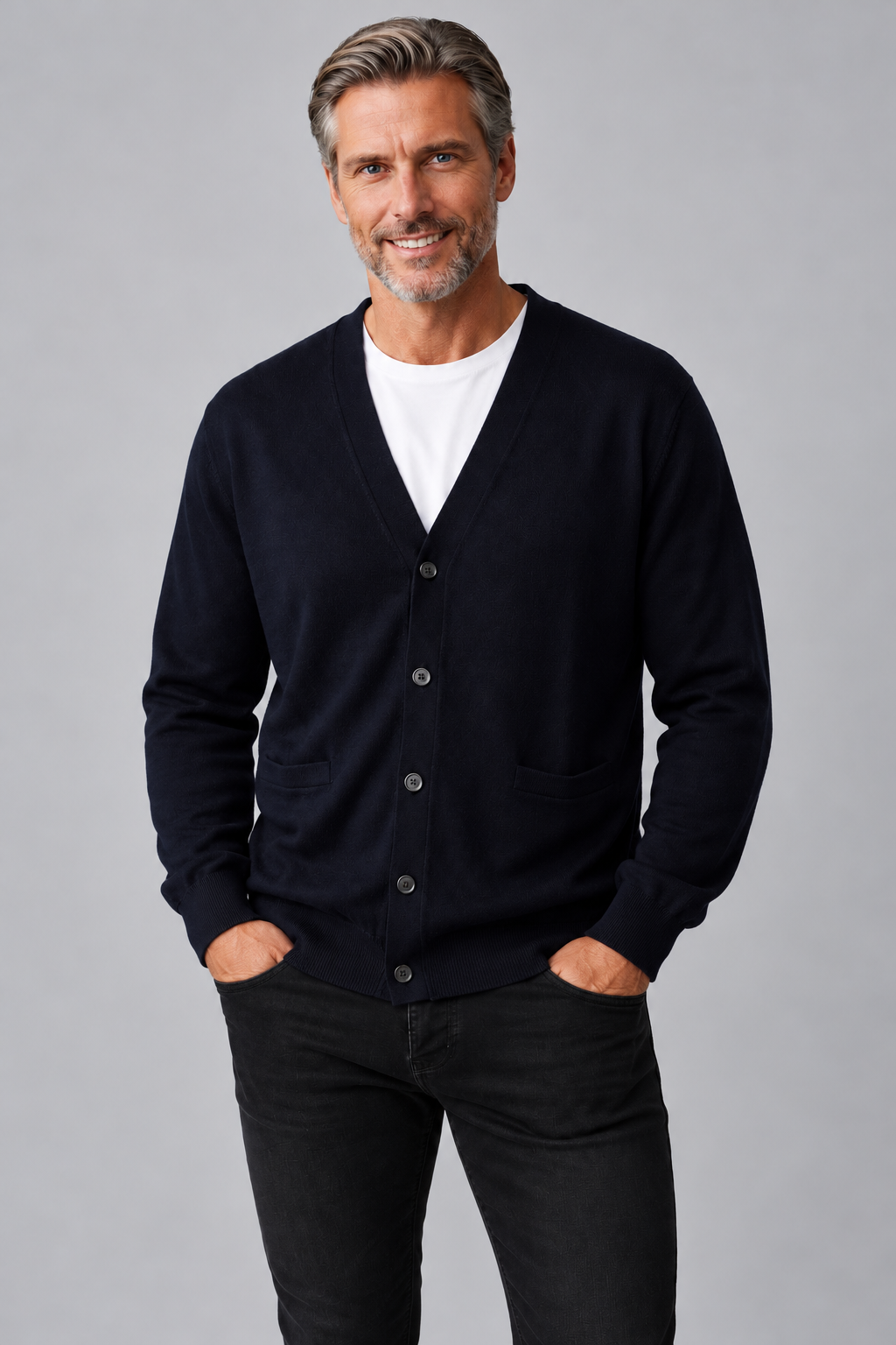 A smiling man with gray hair and a beard wearing a dark navy blue button-up cardigan over a white t-shirt and black jeans, standing against a solid gray studio background.