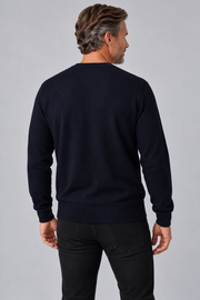 A man viewed from the back wearing a dark navy crewneck sweater and black jeans, standing against a neutral gray studio background.