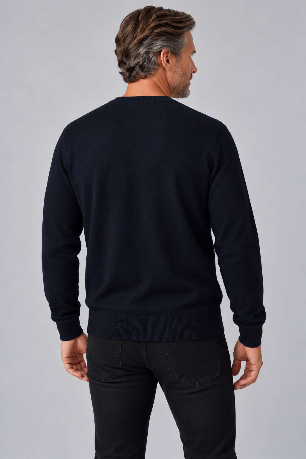 A man viewed from the back wearing a dark navy crewneck sweater and black jeans, standing against a neutral gray studio background.