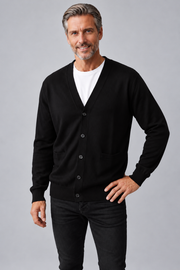 A smiling middle-aged man wearing a black button-front cardigan over a white t-shirt and black jeans, standing against a solid gray studio background.