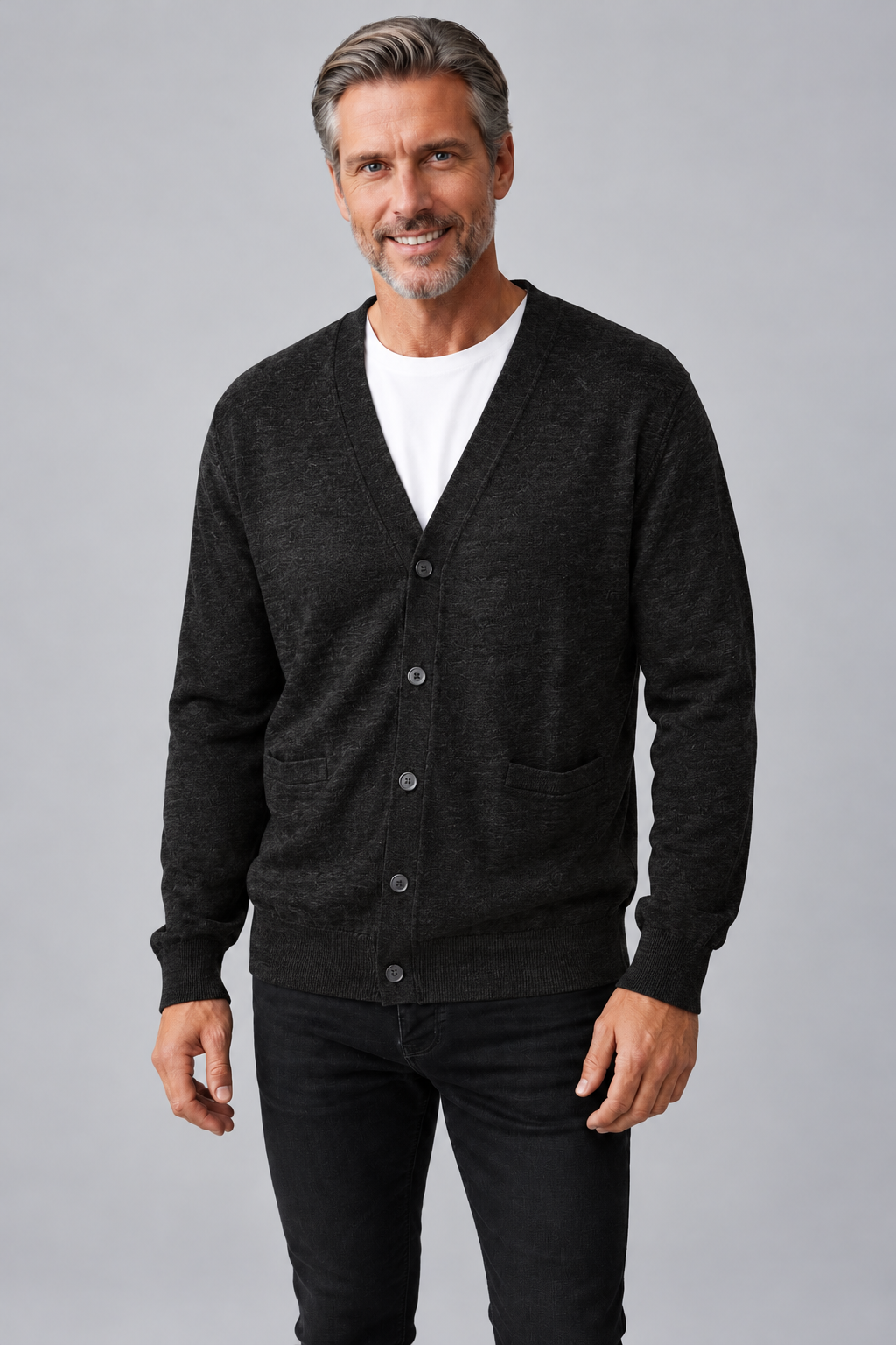 A smiling middle-aged man wearing a dark charcoal gray knit cardigan over a white t-shirt and black jeans, standing against a solid gray studio background.