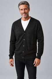 A smiling middle-aged man wearing a dark charcoal gray knit cardigan over a white t-shirt and black jeans, standing against a solid gray studio background.