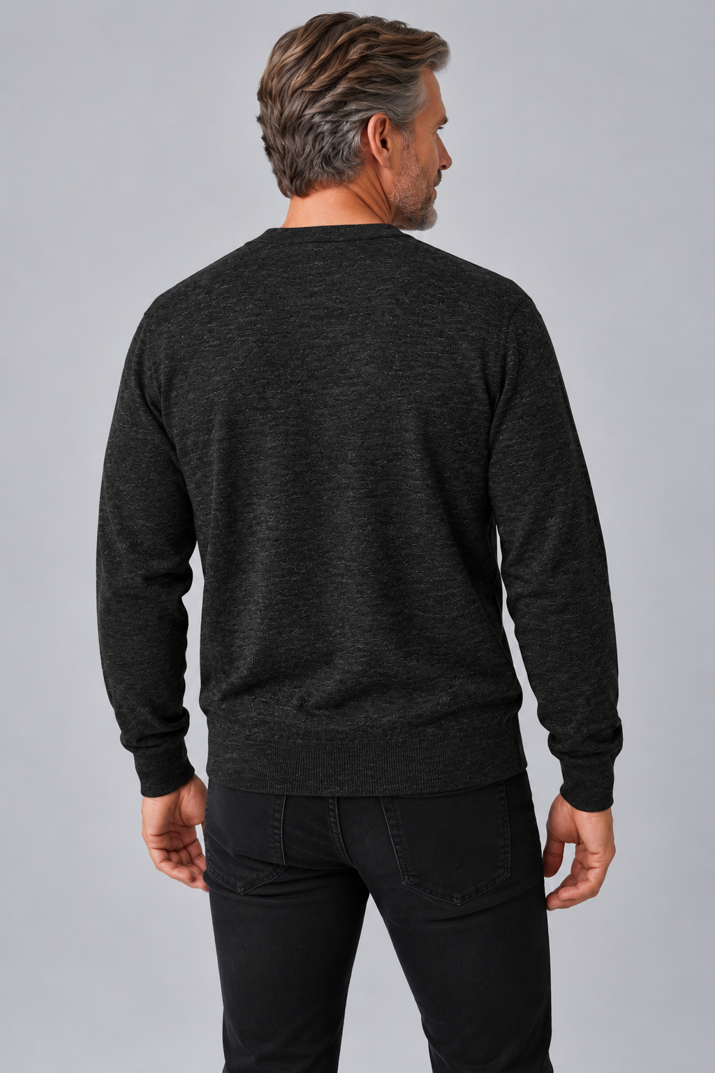 A man viewed from the back wearing a dark charcoal gray long-sleeve crewneck sweater and black jeans, standing against a solid gray studio background.