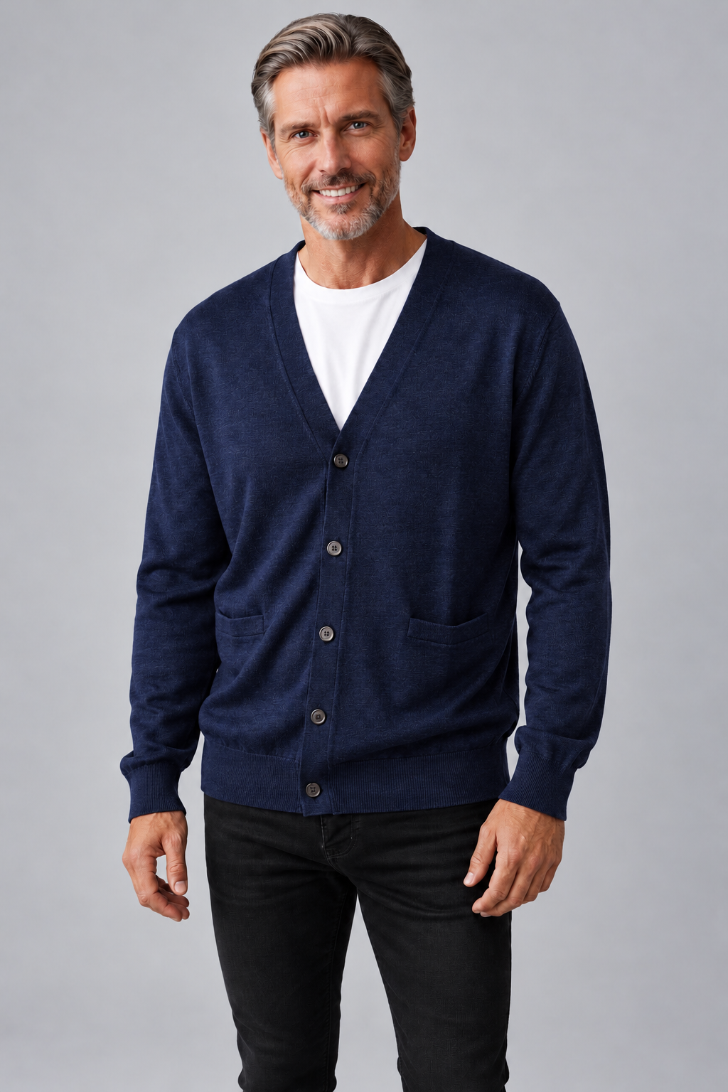 A smiling man wearing a navy blue knit cardigan over a white t-shirt and black jeans, standing against a solid gray studio background.