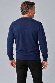 A man viewed from the back wearing a navy blue long-sleeve crewneck sweater and black pants, standing against a solid gray studio background.