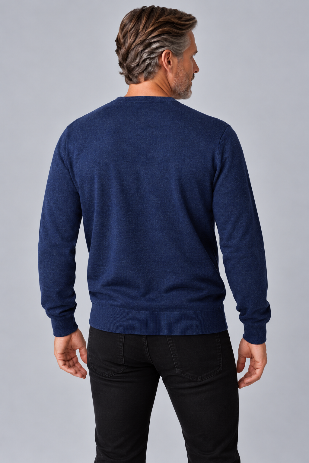 A man viewed from the back wearing a navy blue long-sleeve crewneck sweater and black pants, standing against a solid gray studio background.