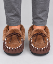 A pair of brown kangaroo fur moccasin slippers with white contrast stitching and dark brown laces, shown on a person's feet wearing grey pants against a light grey background.