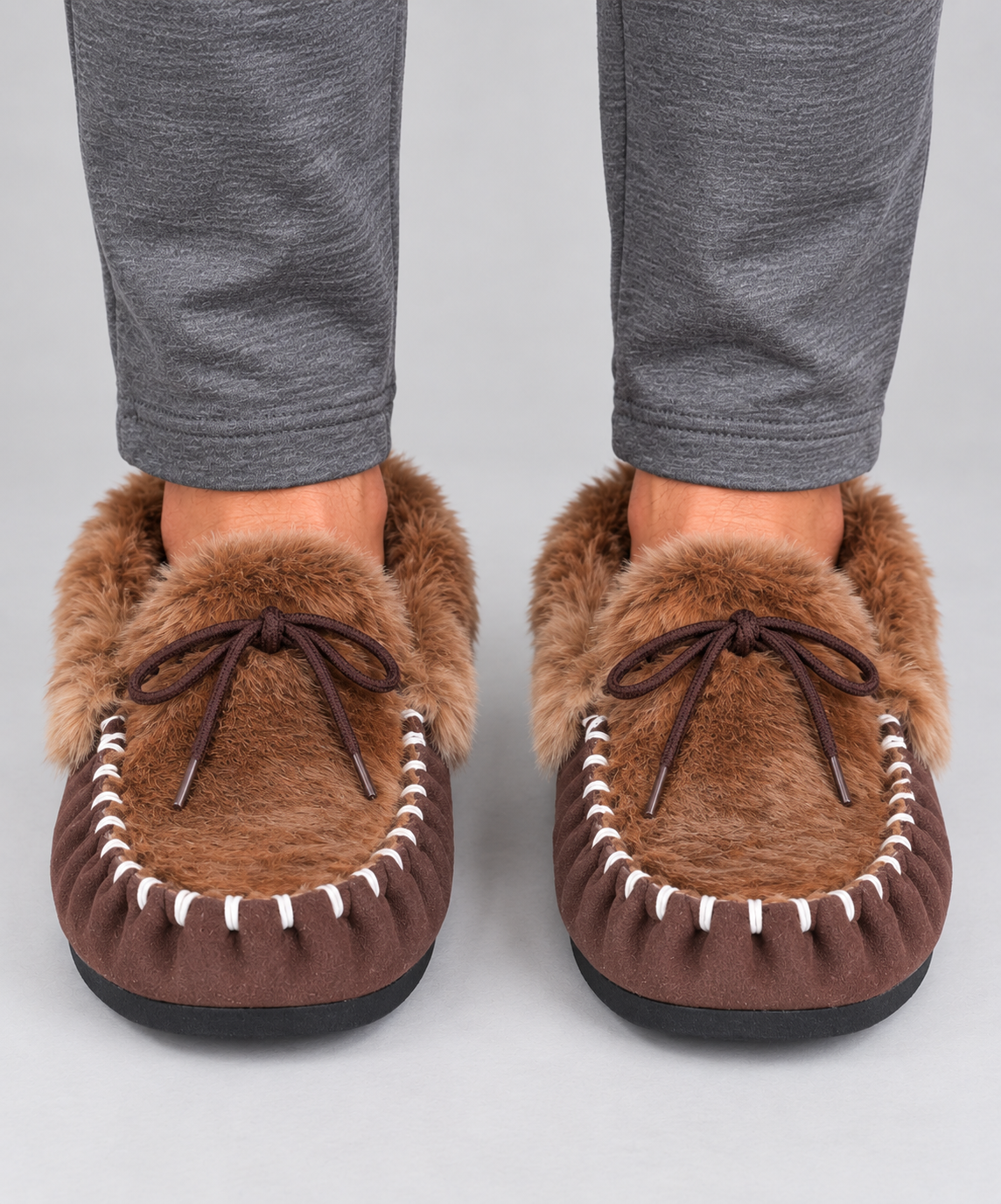 A pair of brown kangaroo fur moccasin slippers with white contrast stitching and dark brown laces, shown on a person's feet wearing grey pants against a light grey background.