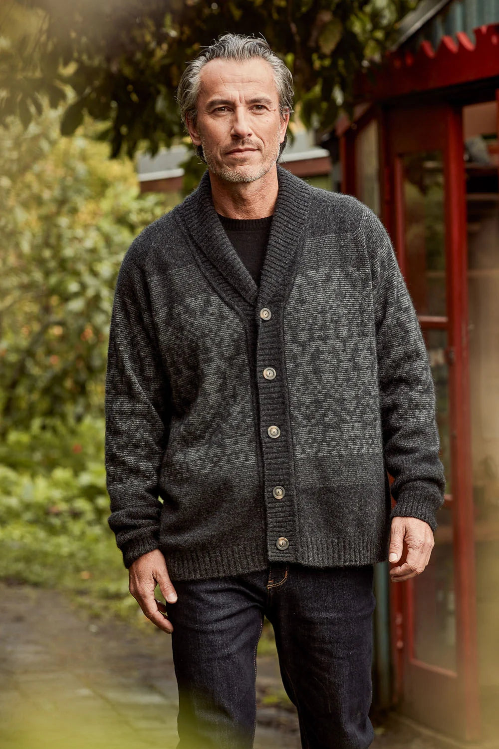 A man wearing a dark gray patterned knit cardigan over a black shirt and dark jeans, standing outdoors in front of a red telephone booth.