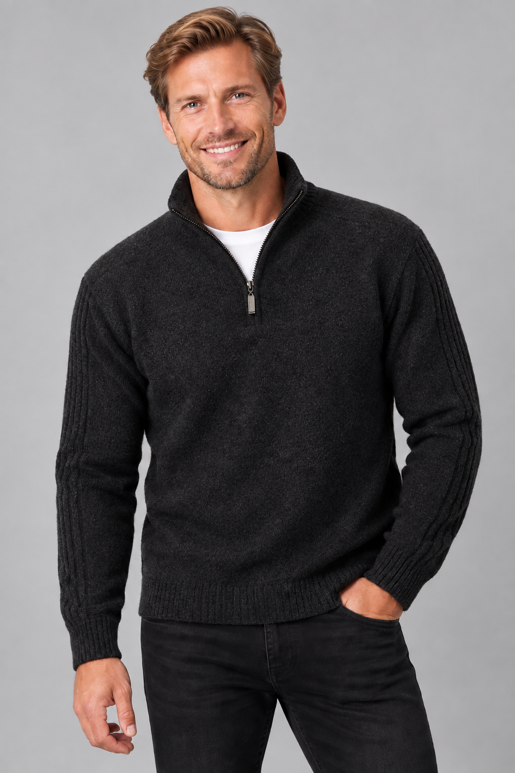 A smiling man wearing a dark charcoal gray quarter-zip knit sweater over a white undershirt and black jeans, standing against a solid gray studio background.
