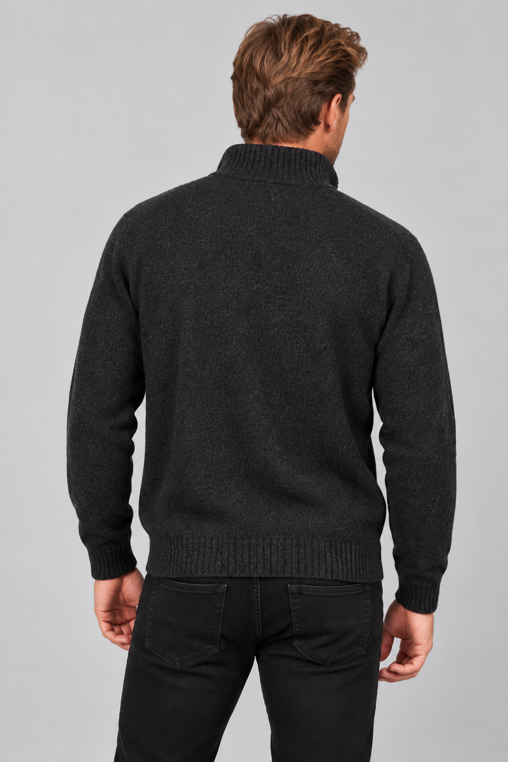 A man viewed from behind wearing a dark charcoal gray ribbed knit sweater and black pants, against a solid light gray studio background.