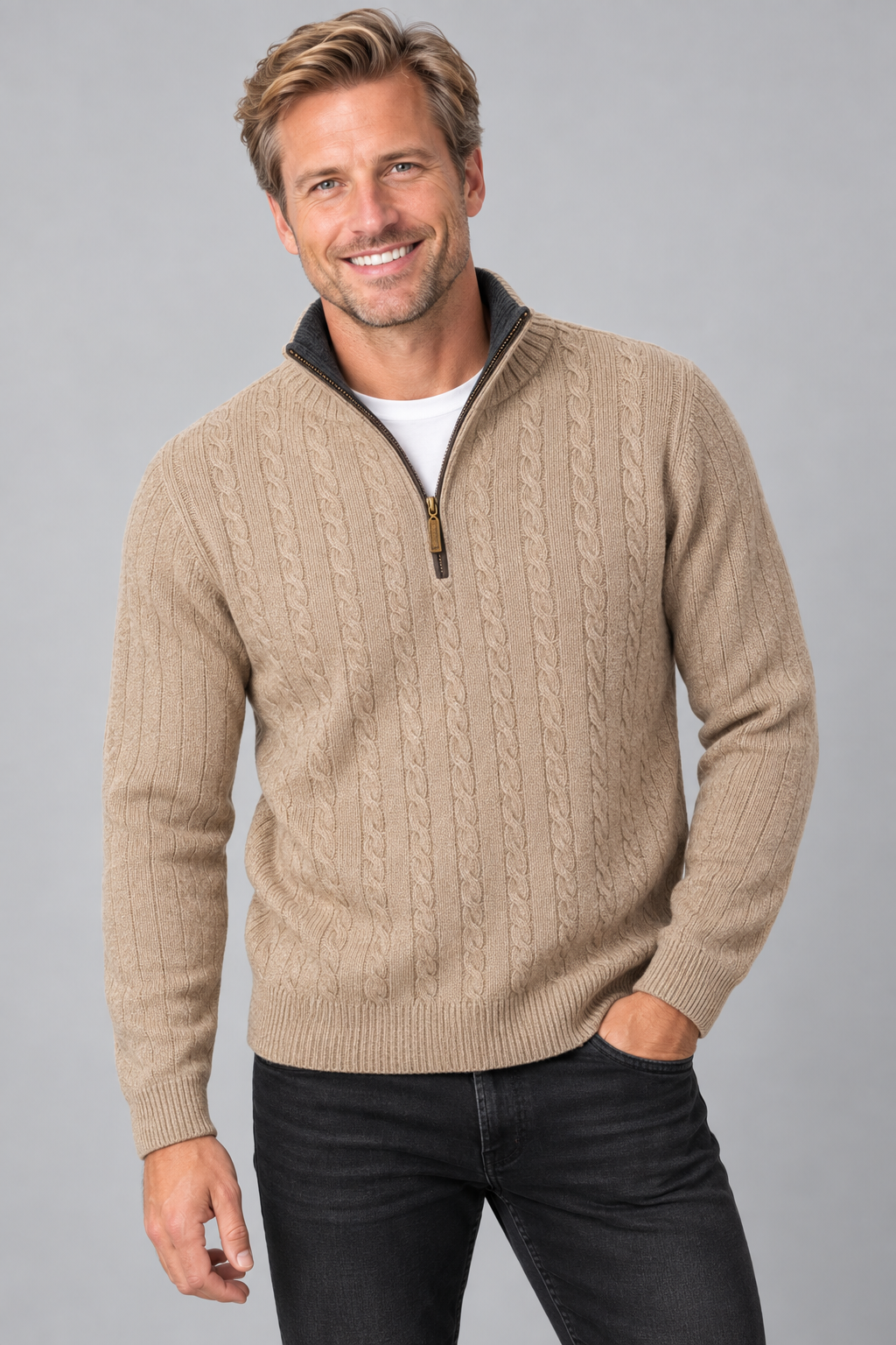 A smiling man wearing a beige cable-knit quarter-zip sweater with a dark collar, paired with a white t-shirt and black jeans, against a solid gray studio background.