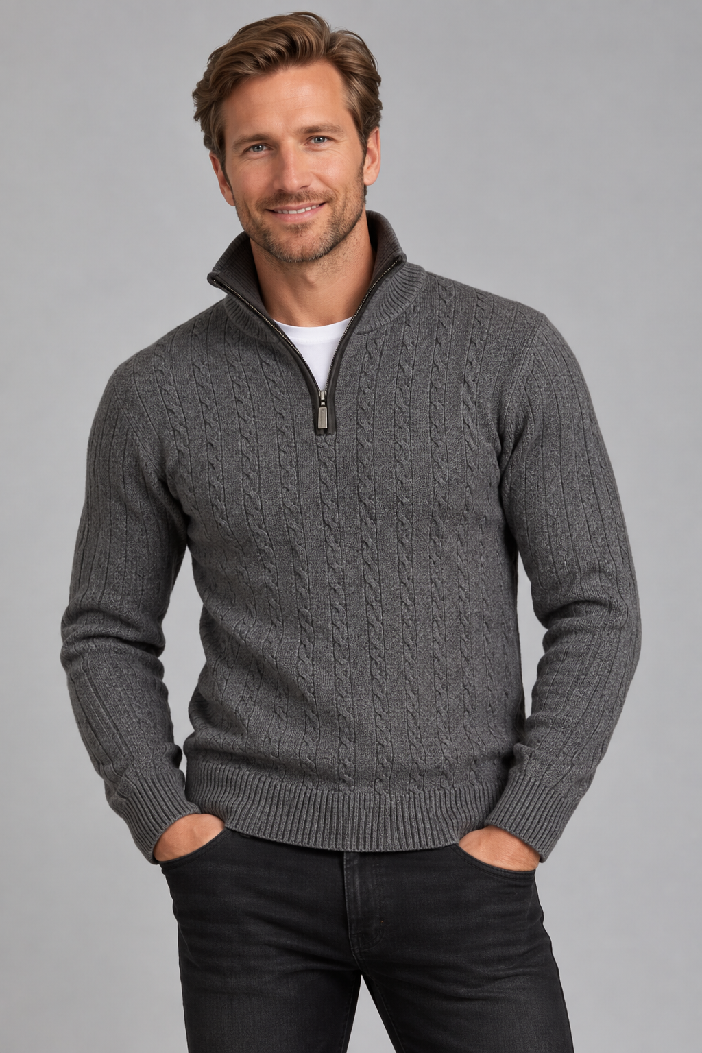 A man wearing a dark gray cable-knit quarter-zip sweater over a white t-shirt and black jeans, standing against a solid gray studio background.