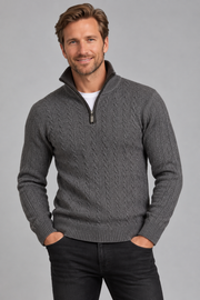 A man wearing a dark gray cable-knit quarter-zip sweater over a white t-shirt and black jeans, standing against a solid gray studio background.