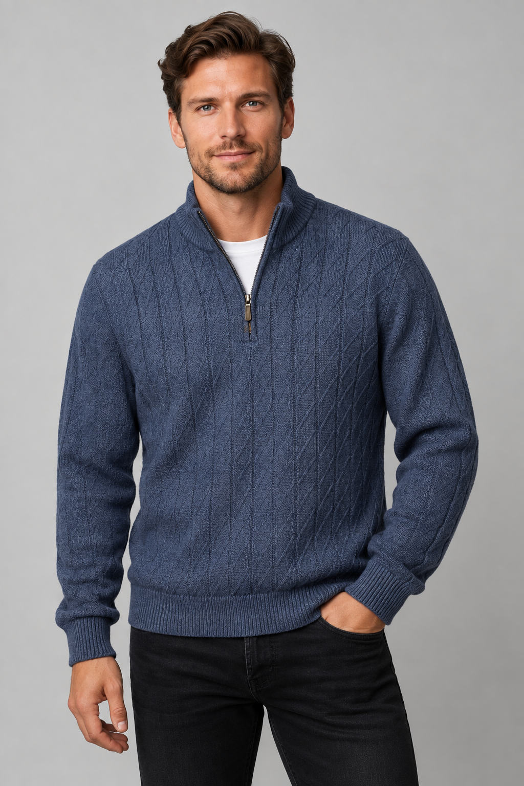 A man wearing a blue cable-knit quarter-zip sweater over a white t-shirt and black jeans, standing against a neutral gray studio background.