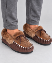 A pair of brown moccasin-style slippers with tan kangaroo fur trim and white whipstitching, shown on a person wearing gray sweatpants against a light gray studio background.